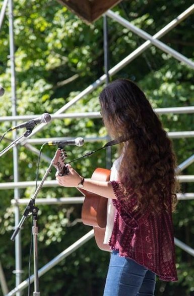 Image Description: Lily Mae playing guitar on an outdoor stage, facing away from the camera.